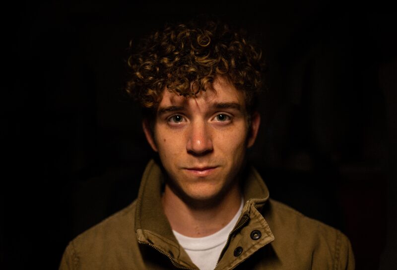 Young man with wistful expression, big dark eyes and thick curly hair in front of a dark background