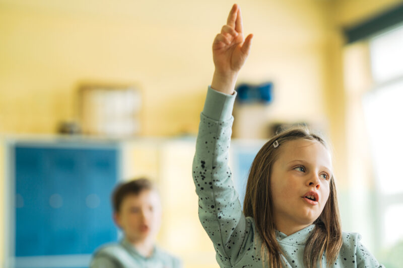 Close-up of a schoolgirl raising hand during lecture in classroom. Elementary school girl student putting hand up for answering the question during lesson.